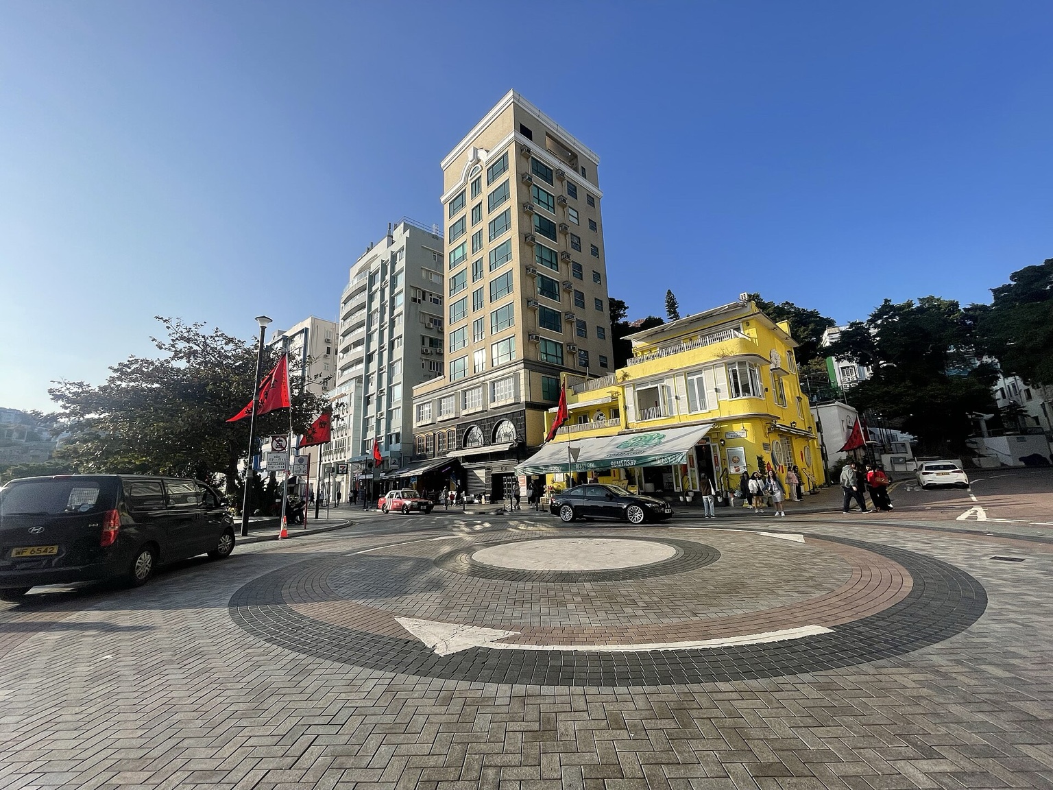香港赤柱主街的真实街区景象，浅色与黄色住宅建筑、路口、树木和行人形成清晰自然的房产生活背景。